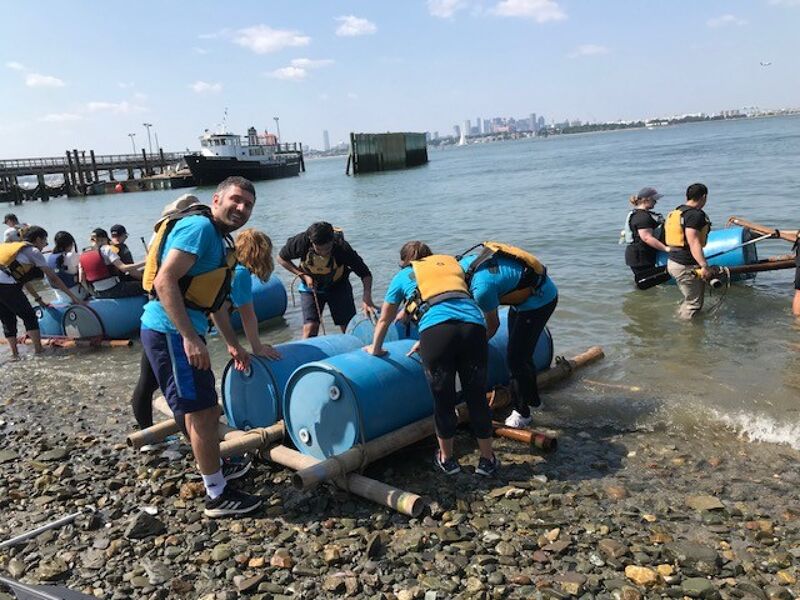 A group of people are working together on a beach to assemble a raft made of blue barrels and bamboo poles. They are wearing life vests, suggesting a water activity or team-building exercise. In the background, a body of water with a distant cityscape and a pier can be seen. The weather appears sunny and pleasant for outdoor activities.